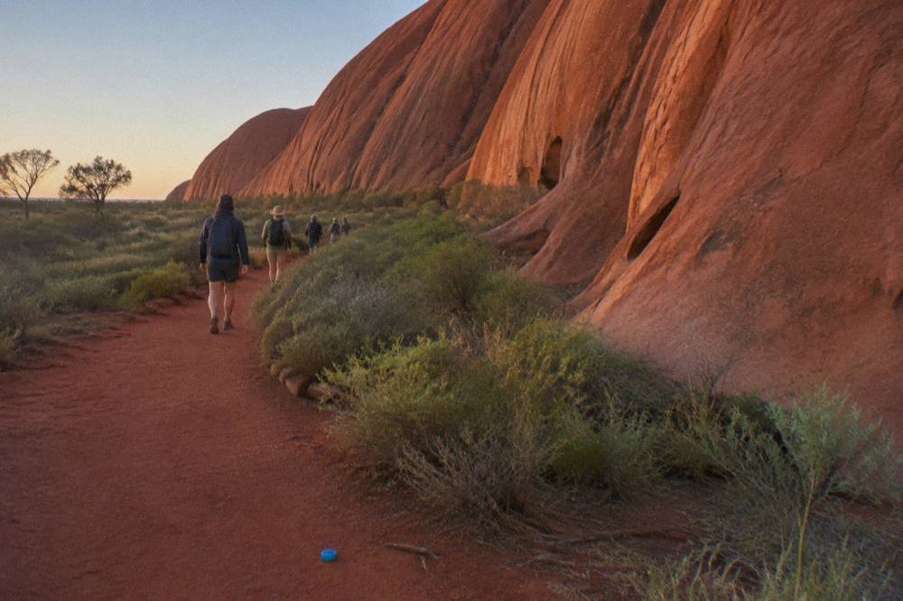A walking track of red earth running close beside the base of Uluru with low shrubs on either side