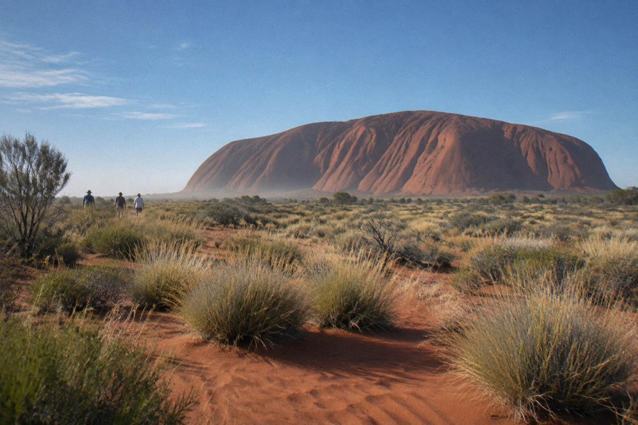 The rust red surface of Uluru rising above sand and spinifex under a wide blue sky