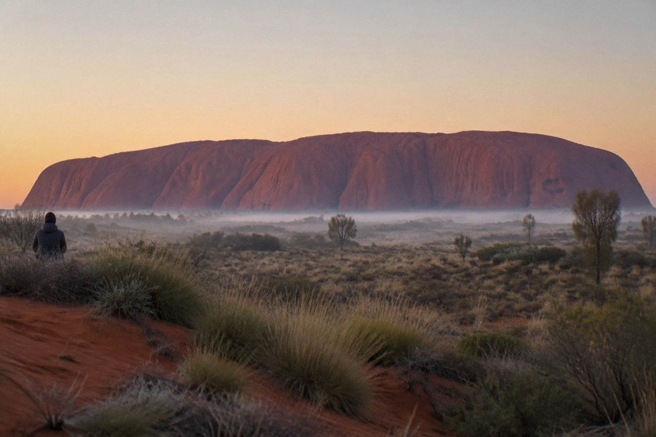 The full length of Uluru seen across red sand plain from a distance at first light