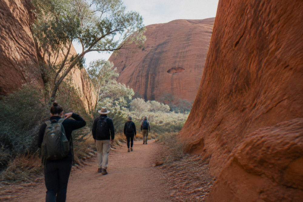 A small group of walkers on a sandy path beside tall orange cliff walls at Uluru