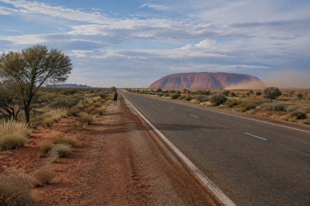 Long straight highway through red desert, distant mountain range, blue sky with clouds