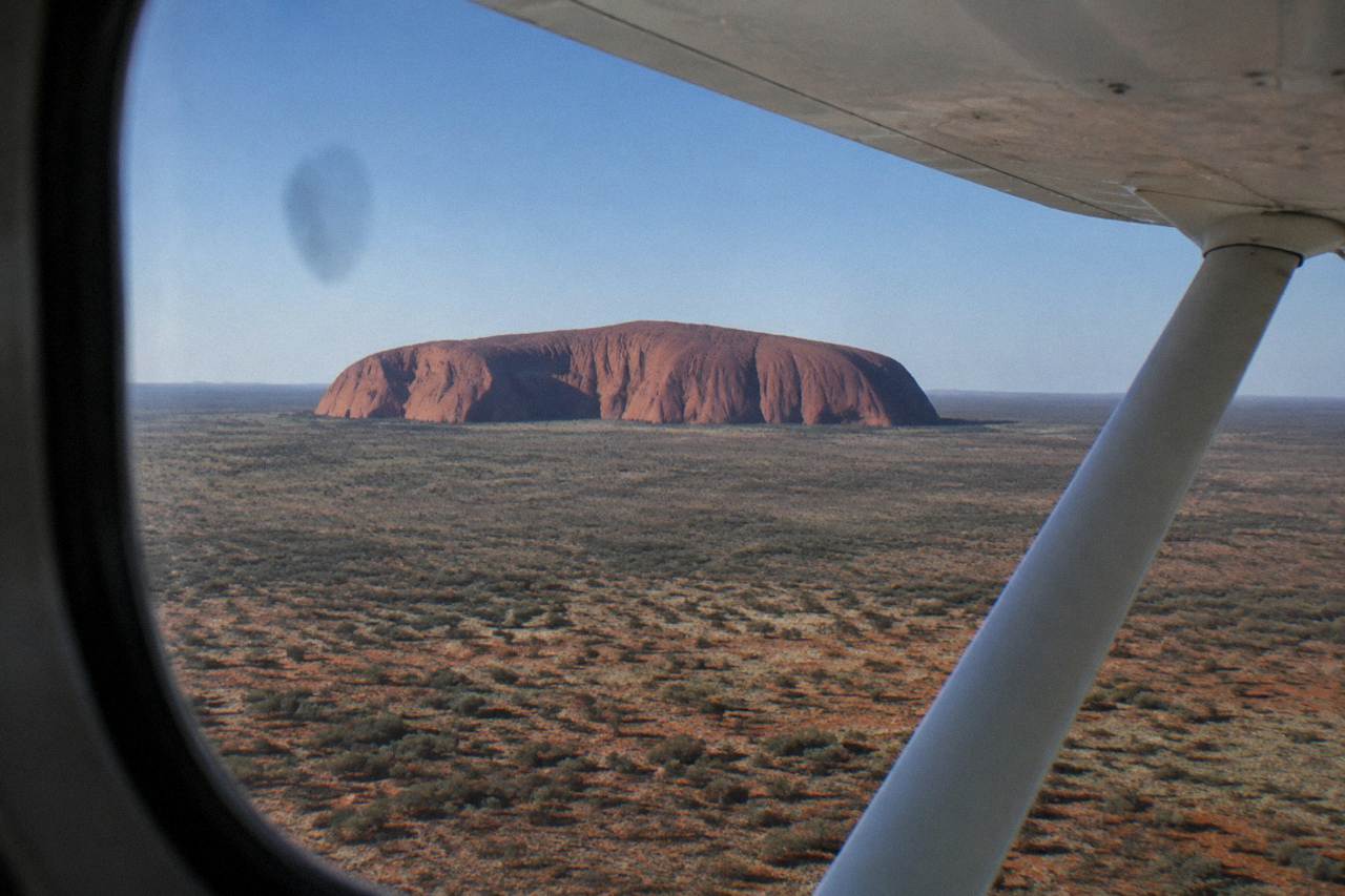 Aerial view of Uluru from a plane window, red desert landscape below, clear sky