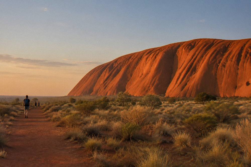 The curved rock face of Uluru glowing orange above sparse desert shrubs in late afternoon light