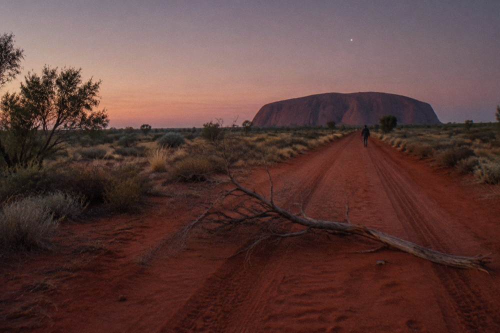 Long red dirt road running straight toward Uluru on the horizon under a wide evening sky