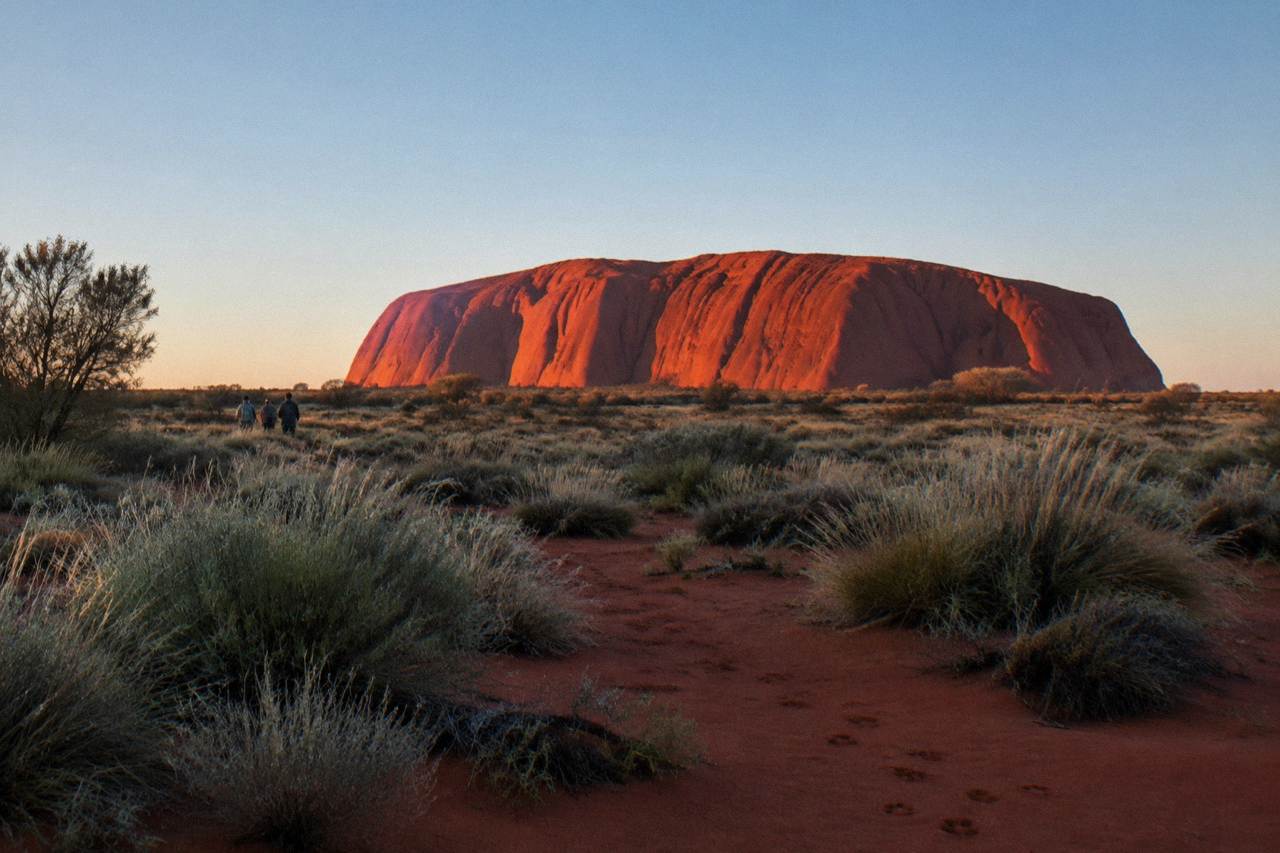 Uluru glowing red under a wide desert sky at dawn with open scrubland in the foreground