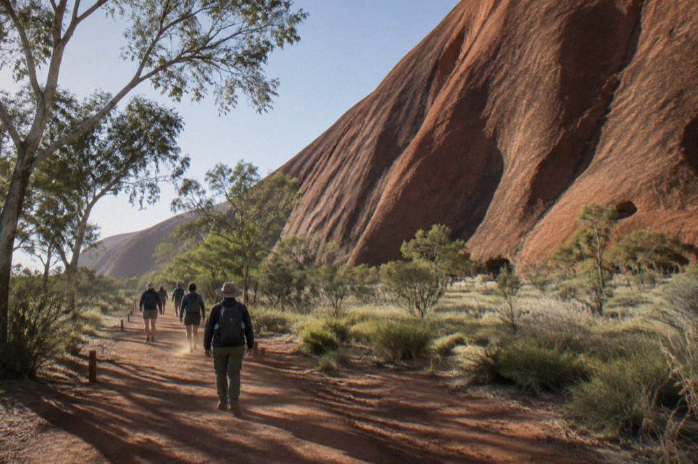 Walkers on a red earth path beside the rock in early morning light, wide desert horizon beyond