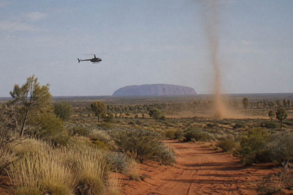A low helicopter flying over red desert country toward the distant shape of Uluru