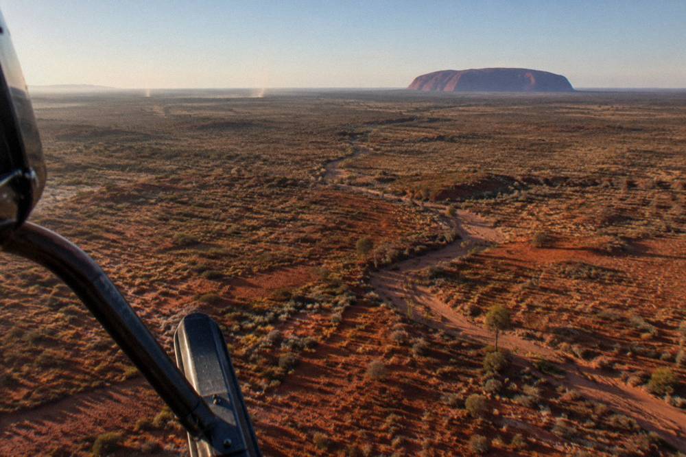 A helicopter flying over red desert country with a large rock formation in the distance