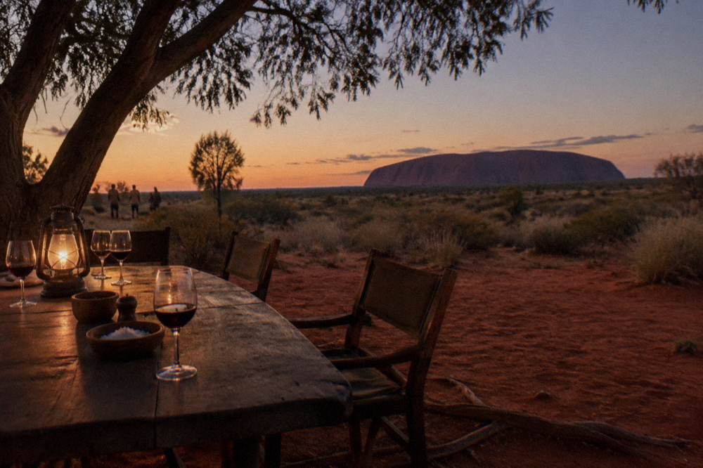 A shaded outdoor dining table set with food and drinks on red desert ground at dusk