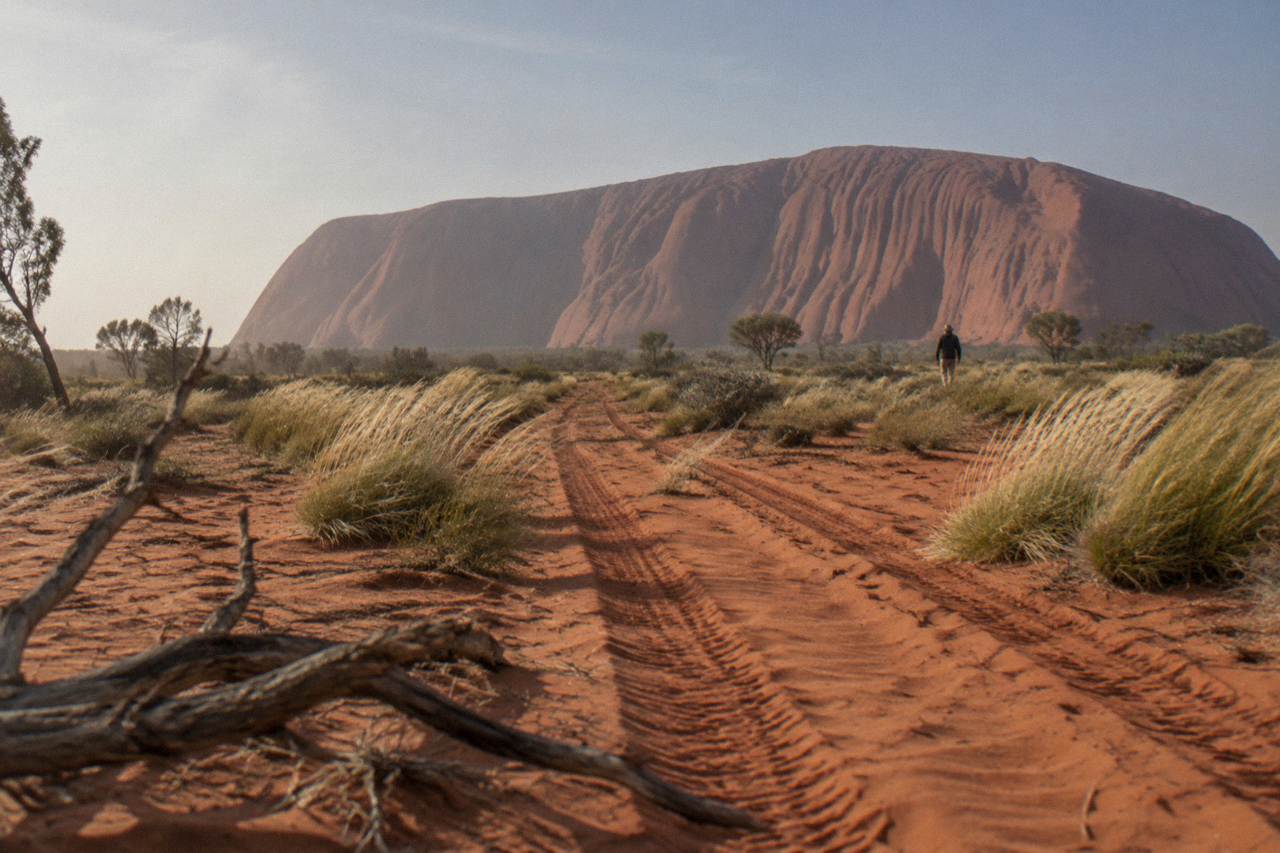 Uluru rising above red desert sand and low scrub under a wide blue sky