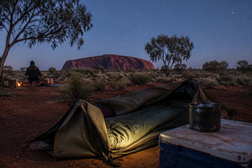 Camping setup with a swag rolled out on red dirt under a clear desert sky