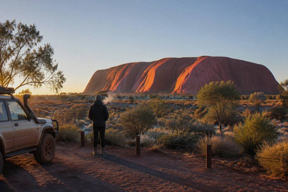 Sunrise light warming the western flank of a large red rock formation above sparse desert ground