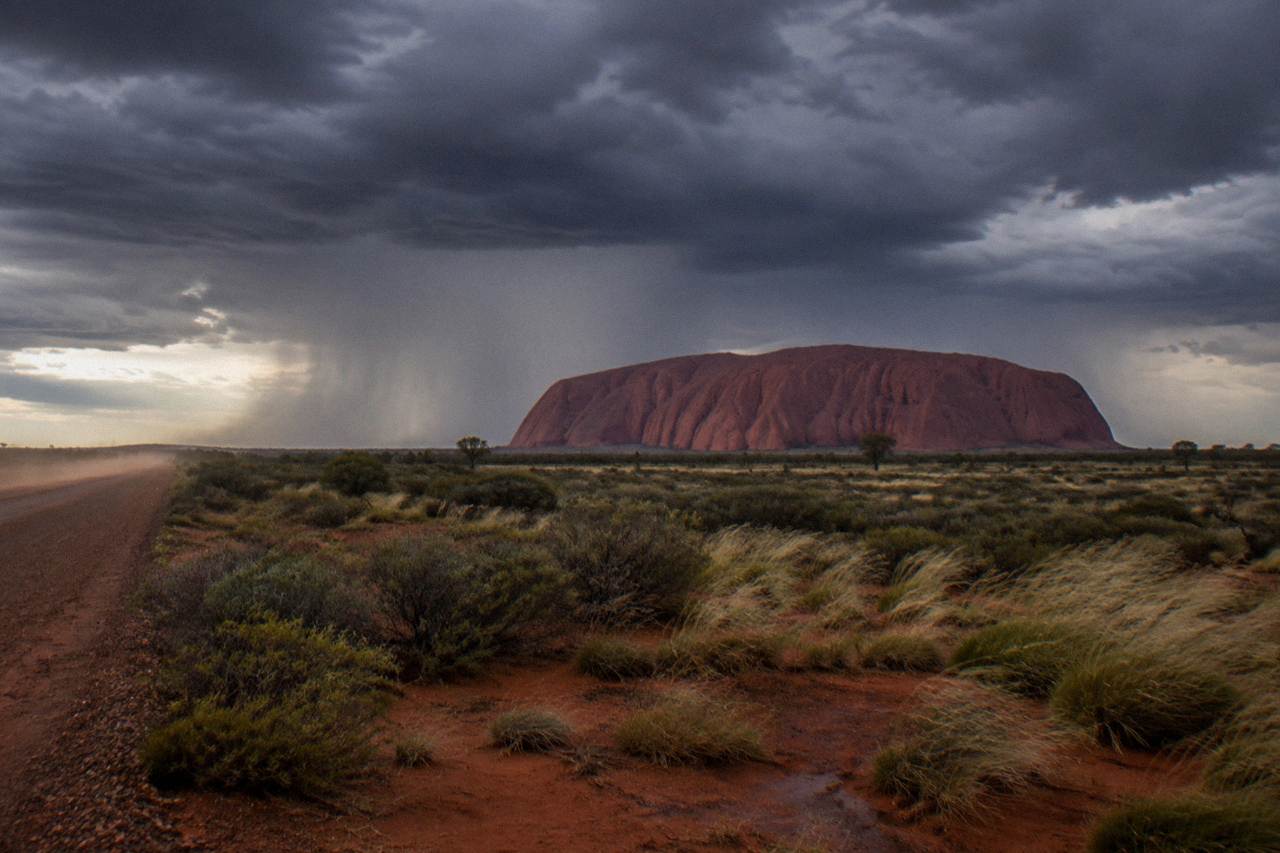 The red sandstone shape of Uluru rising above flat desert scrub under a wide pale sky
