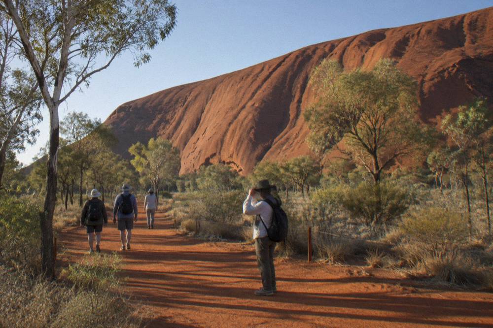 A small group walking on a red dirt path beside desert shrubs in early morning light