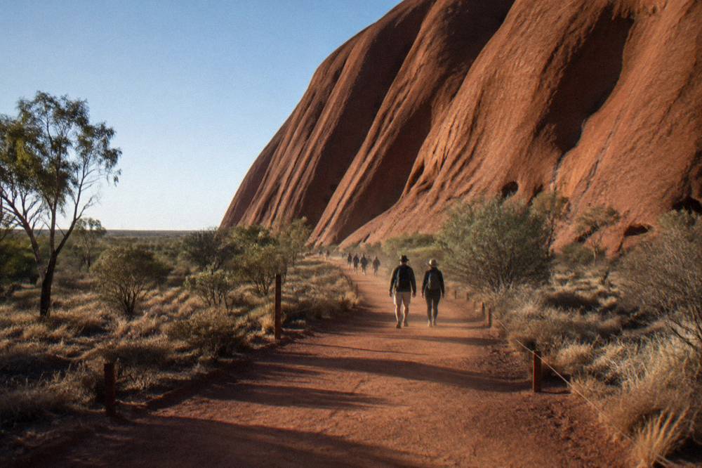 A long desert path curving alongside the sweeping rock wall of Uluru under open sky
