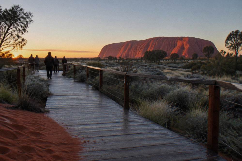 A timber viewing platform facing Uluru as first light warms the desert plain