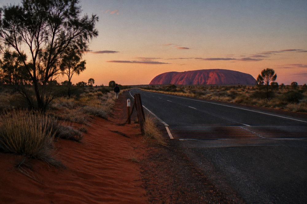 A quiet desert road at first light stretching toward the distant shape of Uluru