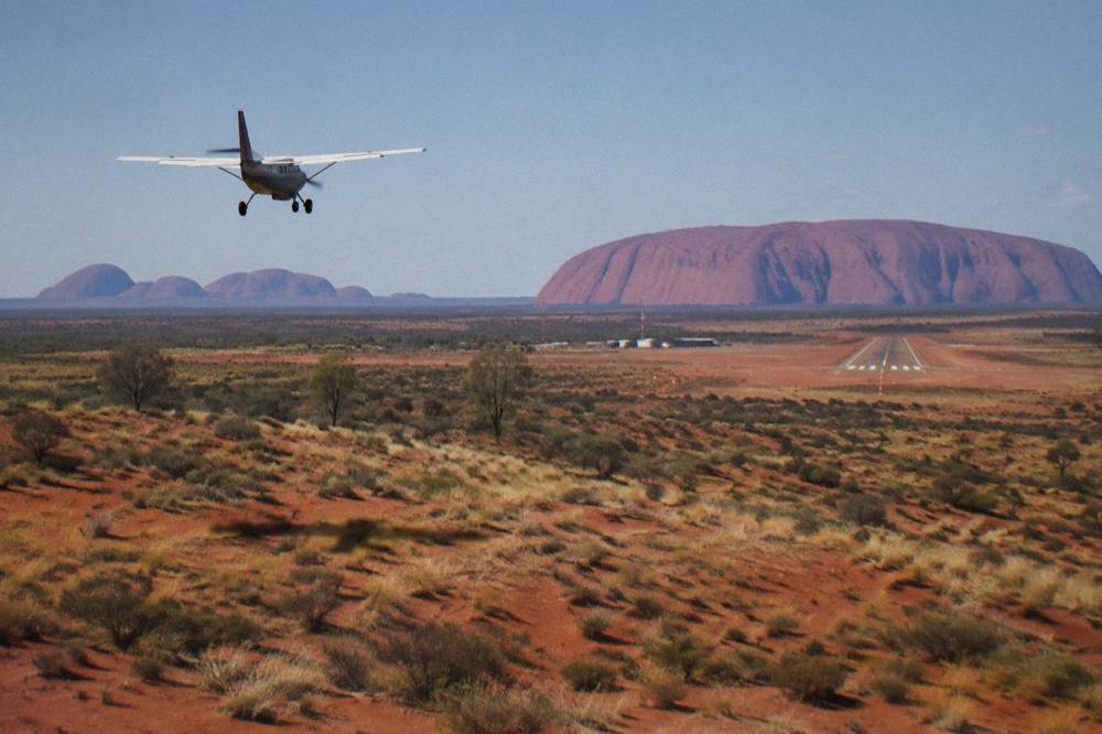 A small aircraft descending over red sand dunes toward a desert airstrip