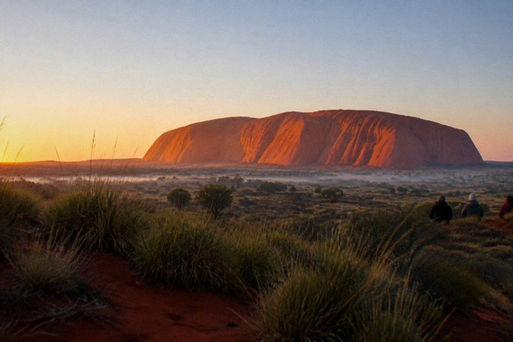 Sunrise light warming the red flanks of Uluru seen from a desert viewing dune