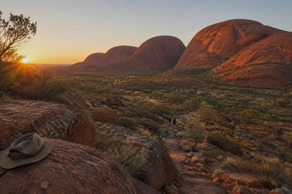 A small group of walkers on a rocky trail below towering orange dome walls in early morning light