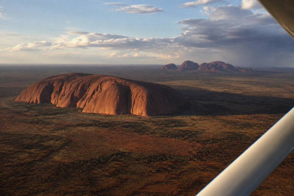 Light aircraft banking over a vast red desert plain toward a single large rock formation