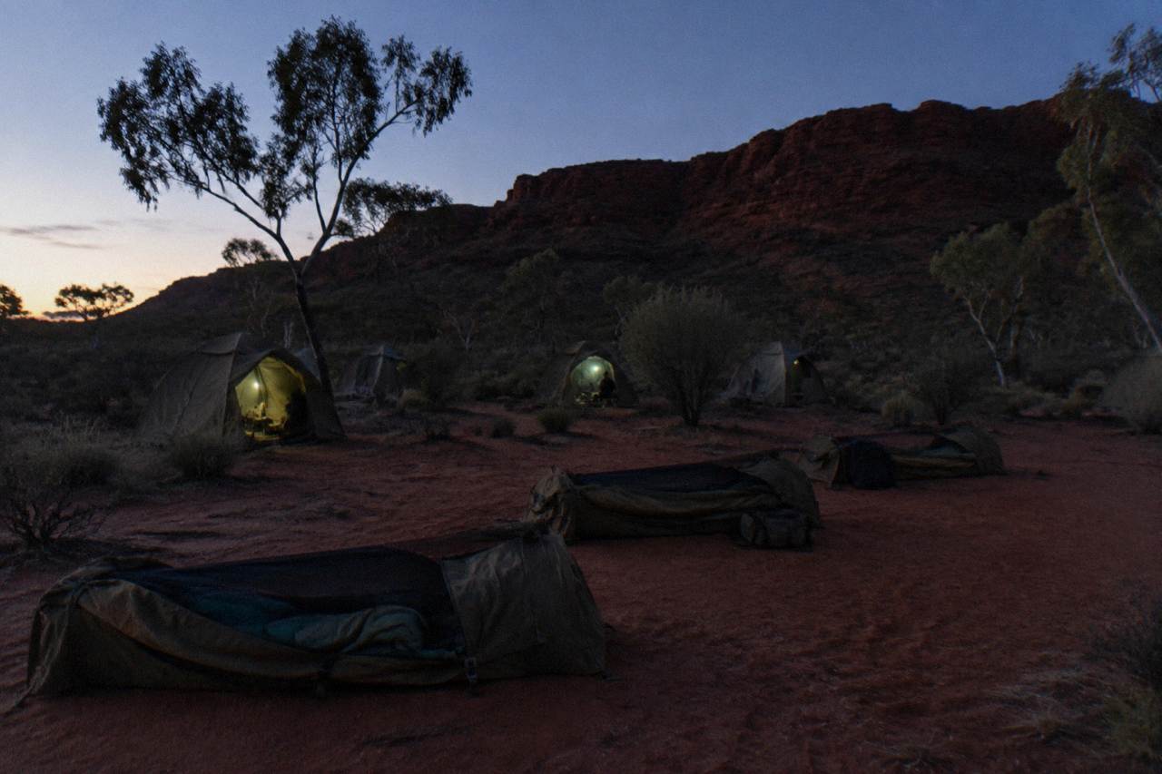 Desert campground at dusk with canvas tents and swags laid out near a low rocky ridge