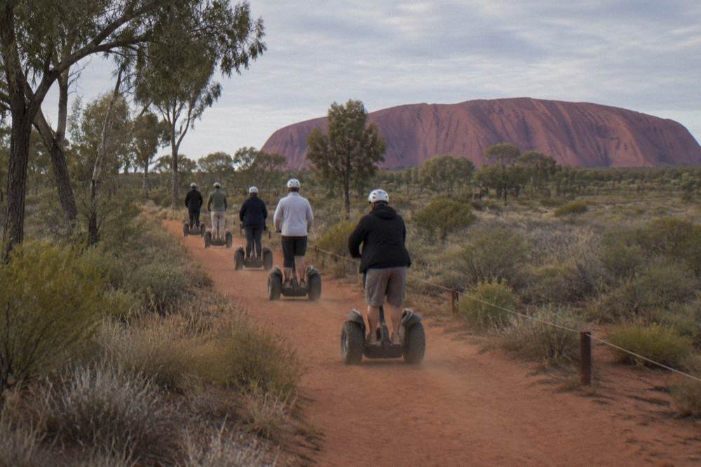 Group of visitors riding Segways in a line along a red dirt path beside desert trees