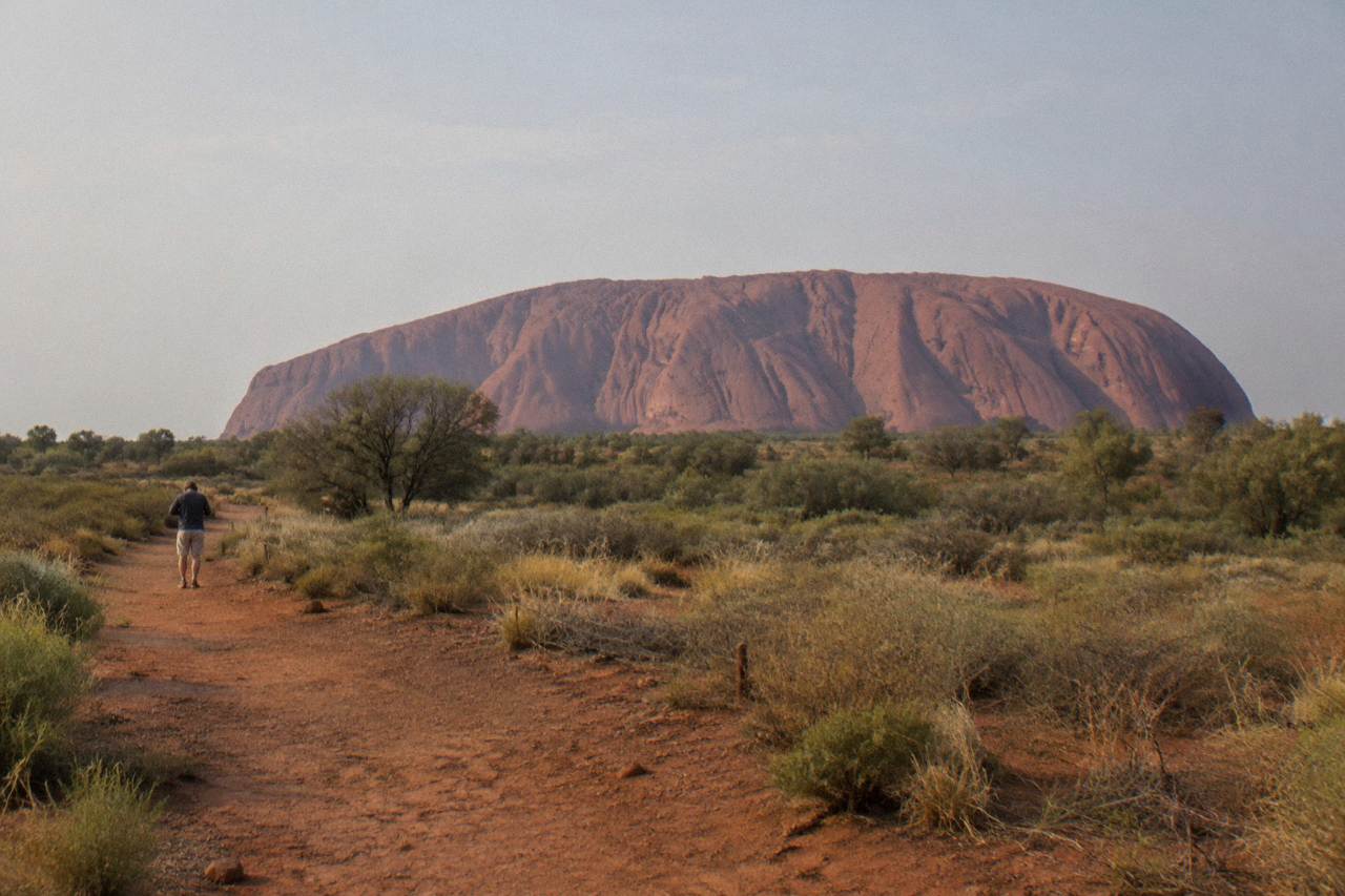 Red rock face of Uluru rising above desert scrub under a pale morning sky