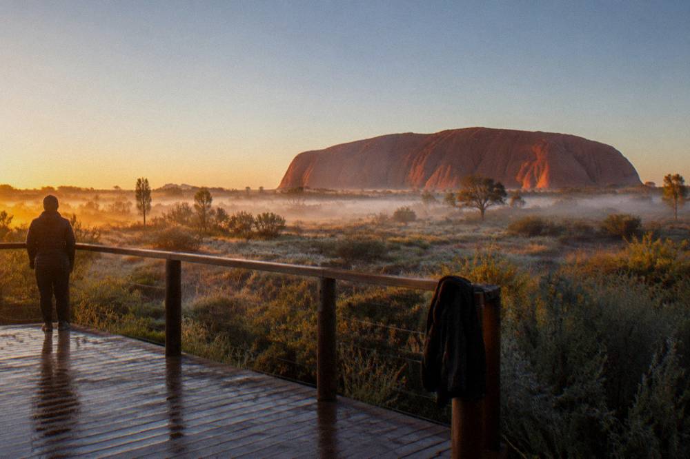 Wooden viewing platform overlooking Uluru glowing red at first light across open scrubland