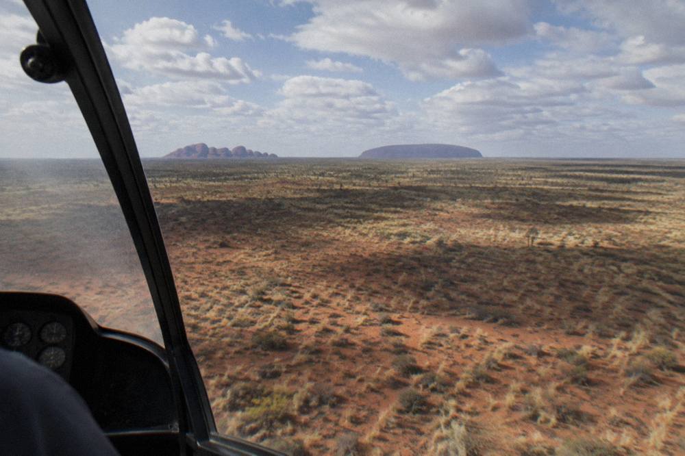 A helicopter cockpit view over red desert plains with the long shape of Uluru on the horizon
