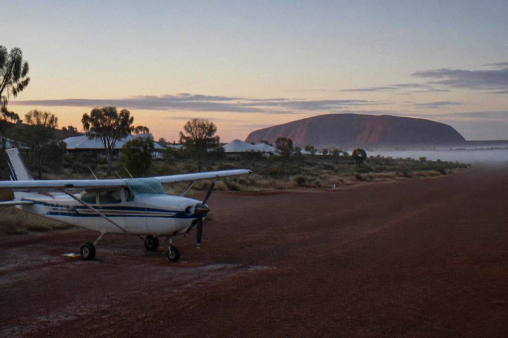 A light aircraft parked on a red dirt airstrip beside the Yulara resort township at first light