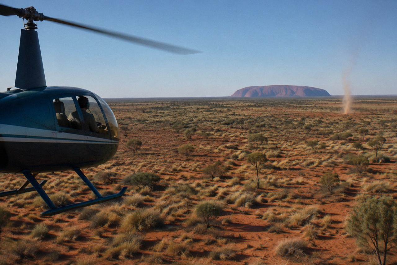 A small helicopter flying low over red desert scrub with Uluru rising in the distance under a wide blue sky