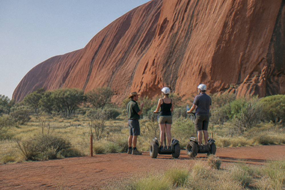 A guide and two riders on Segways pausing beside tall red rock cliffs on a desert path