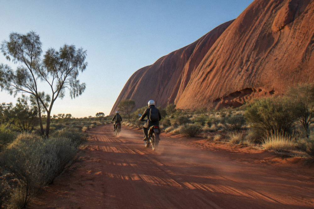 Two riders in helmets on Segways moving along a flat red dirt track beside smooth rock walls
