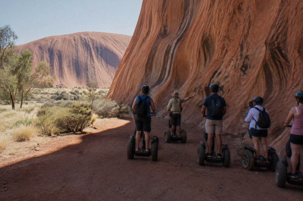 Small group of helmeted riders stopped at a rock face while a guide gestures toward the stone