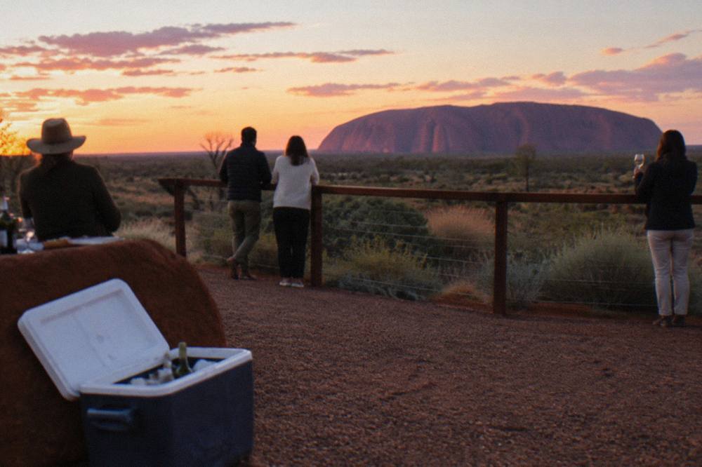 Group of travellers with drinks watching Uluru change colour from a sunset viewing platform