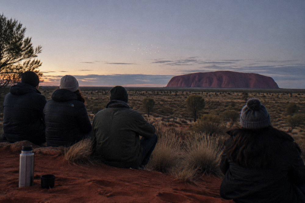 Guests in warm jackets seated on a dark dune watching a light display above the desert